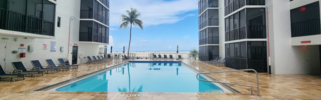 Hotel pool area between white buildings with a palm tree and ocean visible beyond, lined with lounge chairs and umbrellas on a tan tiled deck.